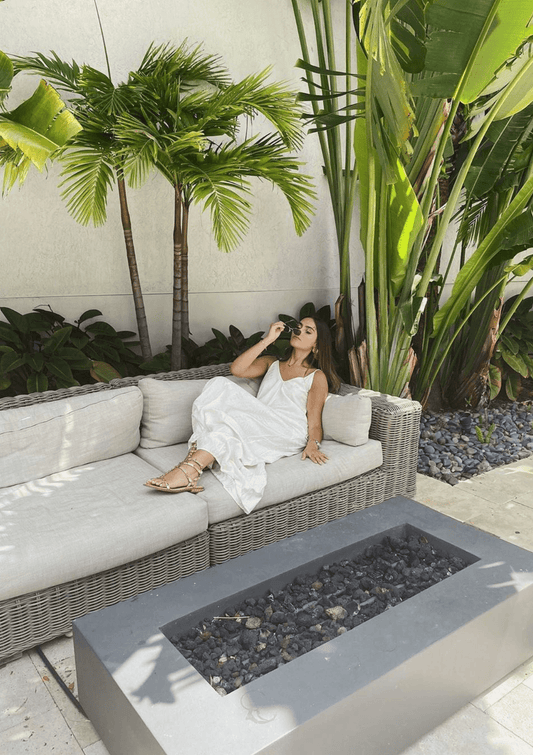 Woman wearing dress outside on wicker patio furniture with palm trees while putting sunglasses on.