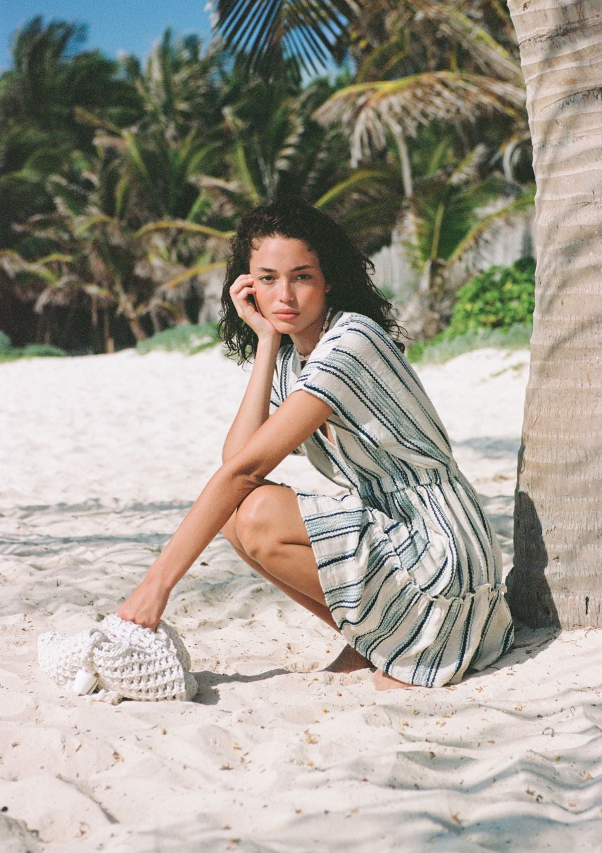 Woman wearing at the ocean beach with handbag in sand and palm trees in back.